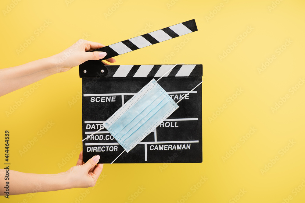 cropped view of camera assistant with clapperboard and medical mask isolated on yellow, cinema concept