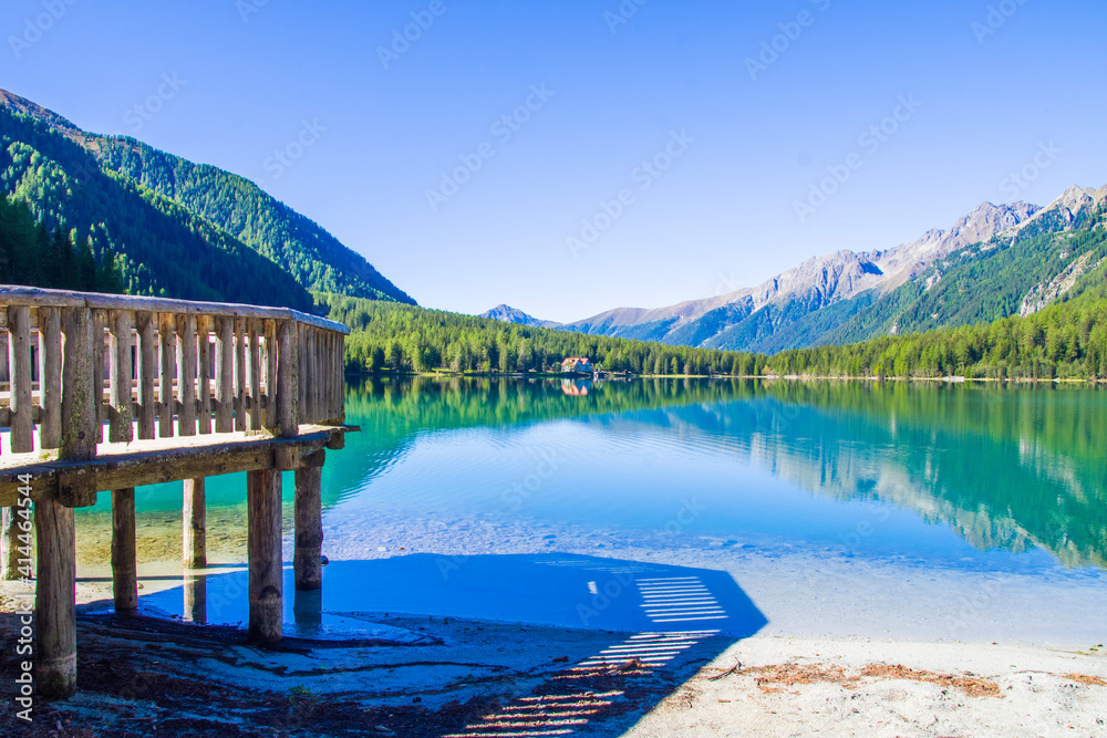 wooden jetty on bright blue mountain lake under blue sky with woods, stones and snow