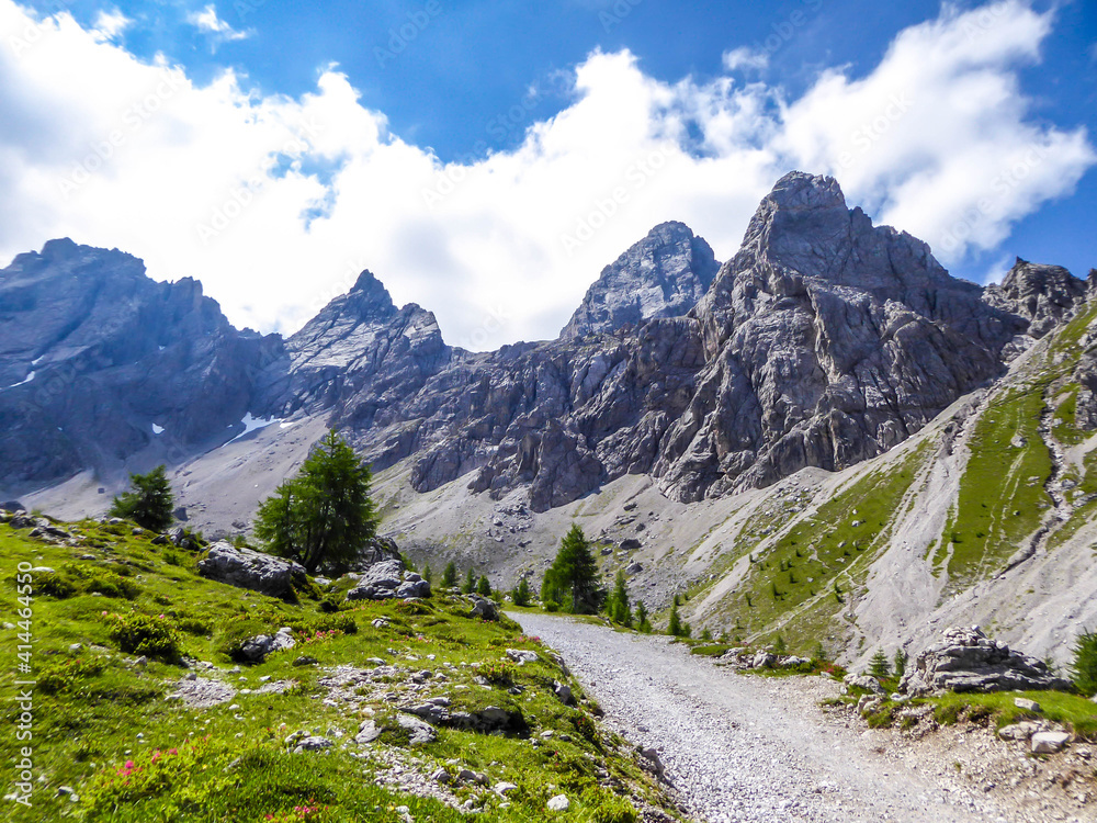 A narrow gravelled road leading to sharp stony mountain range of Lienz Dolomites, Austria. The mountains are partially overgrown with bushes. Dangerous mountain climbing. Tall trees growing in front