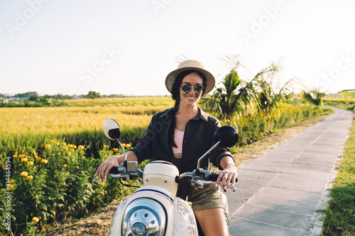 Portrait of joyful female tourist sitting on rented vintage moped smiling at camera while getting to travel destination in Vietnam, funny woman in casual clothing ride on motorcy?le on path photo