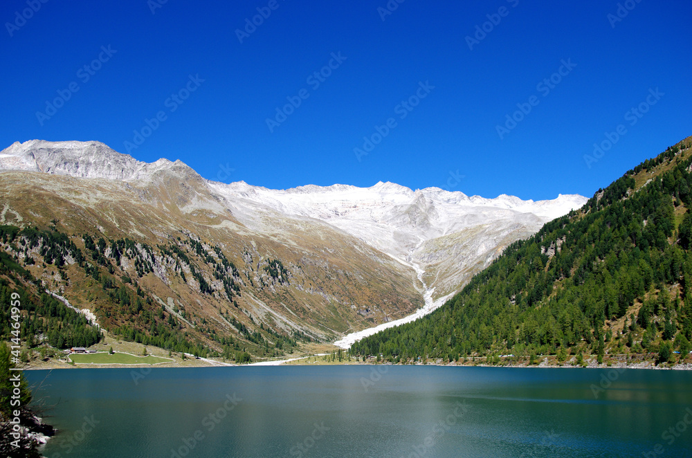 bright blue mountain lake under blue sky with woods, stones and snow