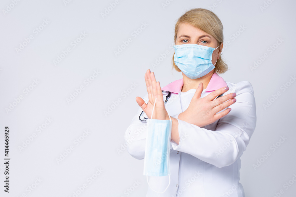 Doctor in protective mask showing stop gesture on gray background. Prevent spreading of coronavirus. Female doctor hands showing crossed stop sign. Health care worker holding medical mask. Doctor in protective mask showing stop gesture on gray background. Prevent spreading of coronavirus. Female doctor hands showing crossed stop sign. Health care worker holding medical mask.