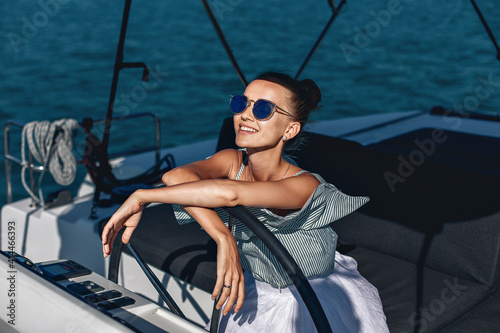 Portrait of smiling happy girl in striped dress and sunglasses sitting at the steering wheel of her own expensive yacht and looking at the road. Laxury concept.