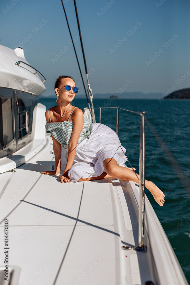 Naklejka premium Portrait of a sexy and charming ladyin a white airy dress and sunglasses sitting on her huge white yacht, sunbathing in the sun and enjoying the local views. Lovely view concept.