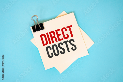 Text DIRECT COSTS on sticky notes with copy space and paper clip isolated on red background.Finance and economics concept.
