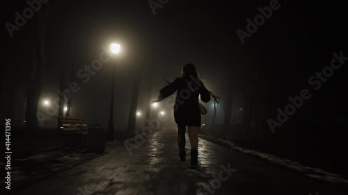 Scared young woman looking back runs away from someone at night in city park photo