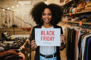 Portrait beautiful young clothing store sales assistant holding "Black friday super sale" sign at business entrance with smile - Owner millennial in shop promotes products and advertises new business