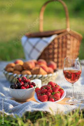 Summertime healthy picnic on nature background at sunny day. Close up Basket fruits strawberries  cherries  glass of red wine  on green grass in garden. Summer weekend outdoor. Beautiful still life