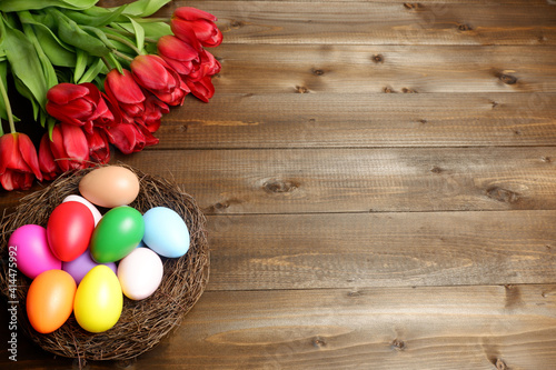 Birds nest with multicolored easter eggs and bouquet of red tulip flowers on dark wooden planks desk. Easter holiday banner  card  poster  header  voucher  invitation template