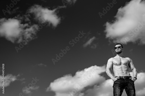 Man with perfect body pumping up muscles with no shirt. Sky with clouds above. Concept of black and white photo.