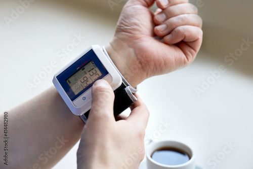 A man measures his blood pressure with a blood pressure monitor. In the background is a coffee cup with coffee. Selective focus.