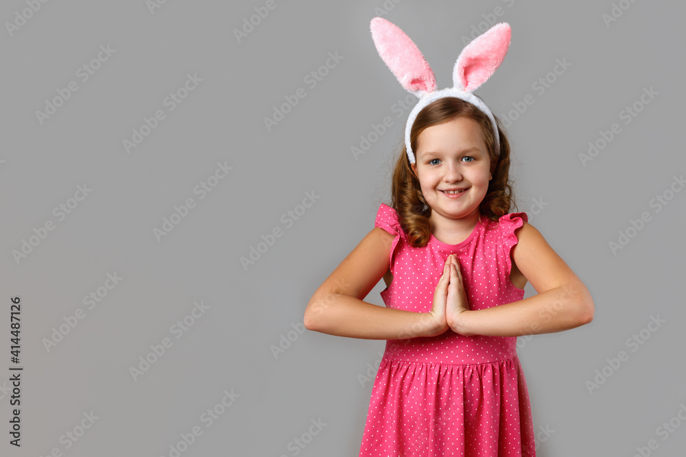 Happy easter. Smiling little girl in a pink dress with polka dots on a gray background. The child in the rabbit ears folded her hands for prayer.