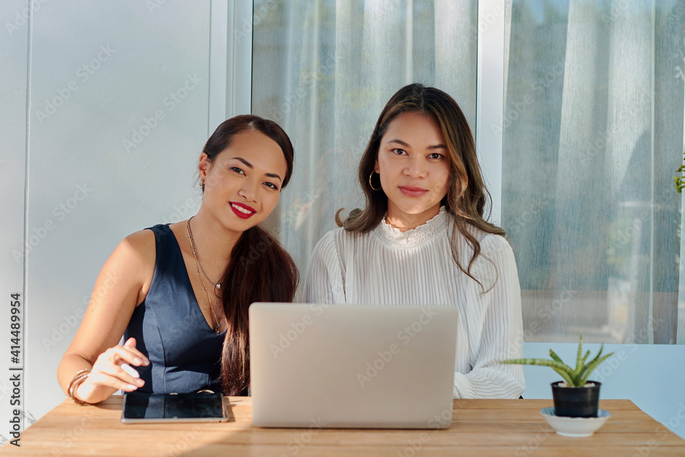 Teamwork of female coworkers outside. Two joyful asian women with laptop pose sitting at table and looking at camera. Teamwork of female coworkers outside. Two joyful asian women with laptop pose sitting at table and looking at camera.