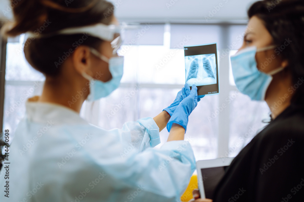 A female doctor in visor and protective gloves discussing an x-ray. Doctor examines an X-ray of a patient s lung infected with covid-19 coronavirus, pneumonia.X-rays of light. Fluorography. A female doctor in visor and protective gloves discussing an x-ray. Doctor examines an X-ray of a patient s lung infected with covid-19 coronavirus, pneumonia.X-rays of light. Fluorography.