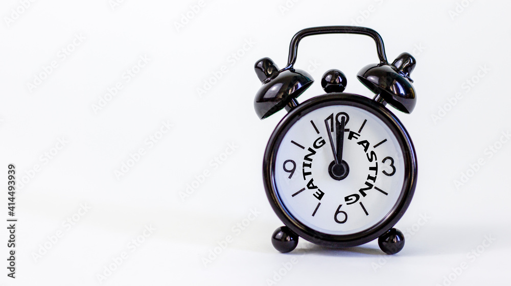 Time to fasting symbol. Beautiful black alarm clock with words 'eating fasting'. Beautiful white background. Copy space. Healthy eating, medical and time to fasting concept. Time to fasting symbol. Beautiful black alarm clock with words 'eating fasting'. Beautiful white background. Copy space. Healthy eating, medical and time to fasting concept.