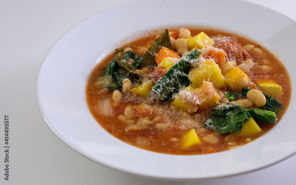 Minestrone soup with beans, butternut squash, kale, bay leaf, and parmesan cheese served on the white shallow bowl. Diagonal view on white background.