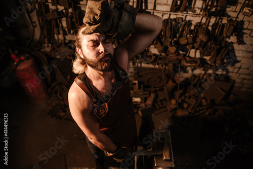 Muscular brutal blacksmith in a leather apron wiping sweat from fatigue in a forge