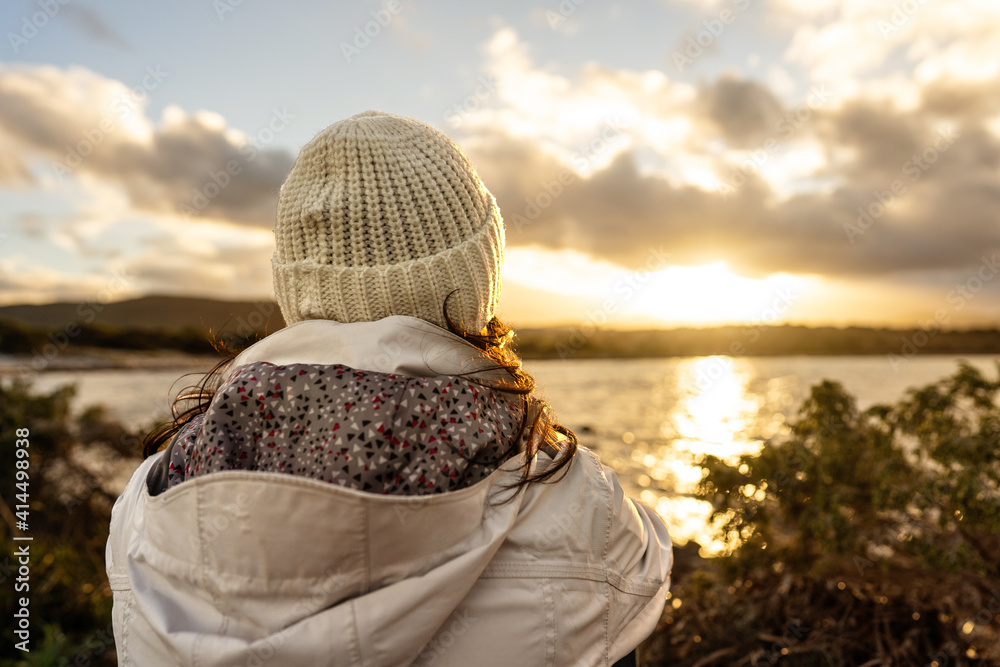 Pensive person view from back looking at the horizon. Unrecognizable woman seen from behind dressed in winter wool hooded clothes looks at a wonderful sunset or sunrise with clouds over the sea Pensive person view from back looking at the horizon. Unrecognizable woman seen from behind dressed in winter wool hooded clothes looks at a wonderful sunset or sunrise with clouds over the sea