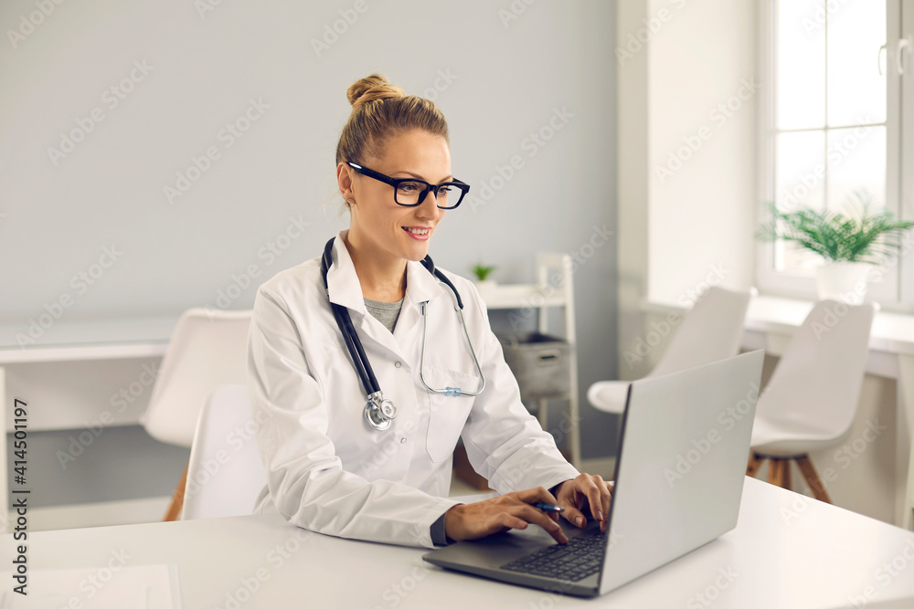 Friendly young doctor sitting at desk, using laptop computer, doing medical Internet research, entering electronic health records into database or giving online consultation to patient on eHealth chat Friendly young doctor sitting at desk, using laptop computer, doing medical Internet research, entering electronic health records into database or giving online consultation to patient on eHealth chat