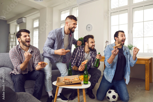 Cheering, shouting young men watching exciting sports game. Group of friends enjoying World Cup on TV at home, eating pizza and chips, drinking beer and celebrating favorite soccer team scoring a goal photo