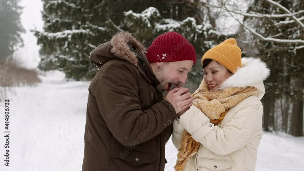 Medium side view of loving Caucasian man warming hands of his Asian wife holding them, breathing on them. Beautiful couple wearing warm clothing, smiling, having date in snowy forest Medium side view of loving Caucasian man warming hands of his Asian wife holding them, breathing on them. Beautiful couple wearing warm clothing, smiling, having date in snowy forest