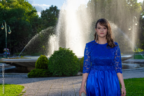 Portrait of young girl with serious look. Beautiful makeup and occasion or evening dress. Adorable fashionable female model posing in the park at fountain in background.