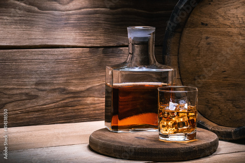 Barrel of scotch whiskey with a decanter of whiskey and a glass of whiskey with ice on a wooden background.