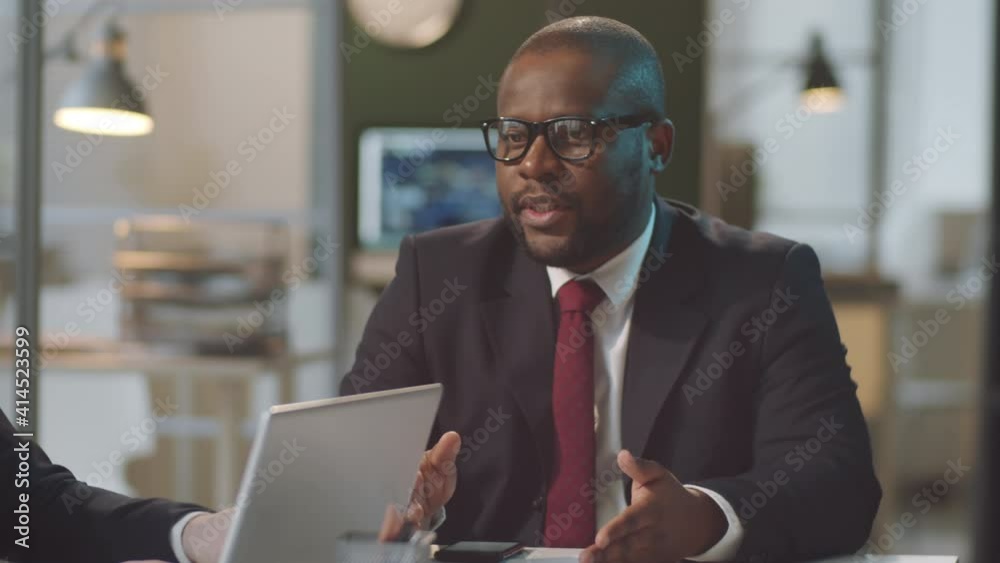 African American and Caucasian business partners in formalwear sitting at office table and discussing project African American and Caucasian business partners in formalwear sitting at office table and discussing project