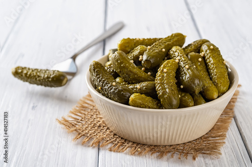 Crunchy pickled cornichons in a beige ceramic bowl and one on a fork on a white wood table. Whole green gherkins marinated with dill and mustard seeds. Delicious baby pickles. Tasty canned vegetables.
