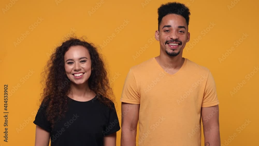 Cheerful smiling young couple two friends african american man woman 20s in basic t-shirts posing isolated on yellow color background studio. People lifestyle concept. Showing ok okay gesture blinking Cheerful smiling young couple two friends african american man woman 20s in basic t-shirts posing isolated on yellow color background studio. People lifestyle concept. Showing ok okay gesture blinking