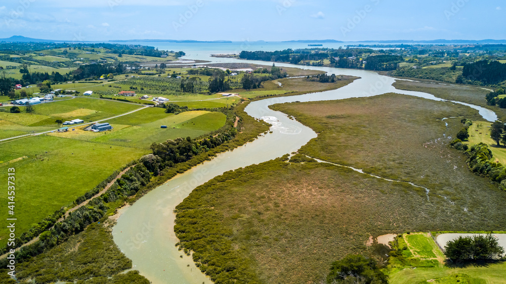 Aerial view of a small river running to the sea through green countryside spotted with little farms. Auckland, New Zealand.