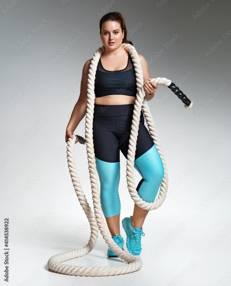 Sporty girl with heavy ropes on her shoulders. Photo of model with curvy figure in fashionable sportswear on grey background. Sports motivation and healthy lifestyle Sporty girl with heavy ropes on her shoulders. Photo of model with curvy figure in fashionable sportswear on grey background. Sports motivation and healthy lifestyle