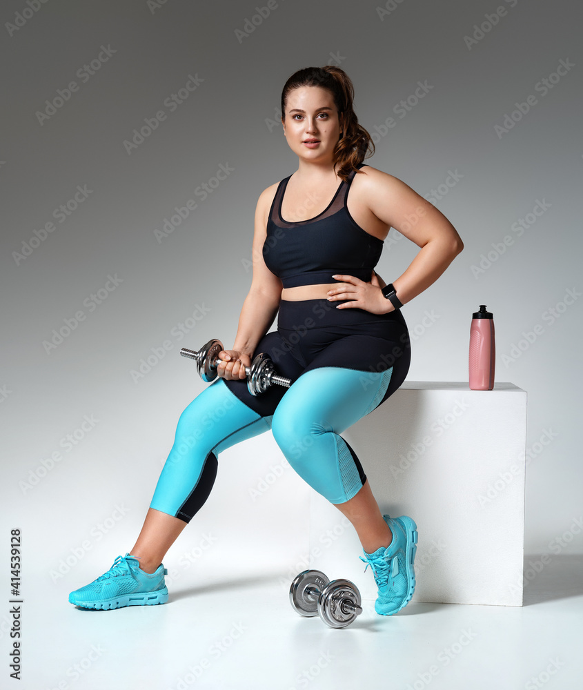 Fototapeta premium Sporty girl resting after workout with dumbbells. Photo of model with curvy figure in fashionable sportswear on grey background. Sports motivation and healthy lifestyle