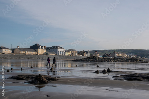 a couple walking along the beach with tree stumps from the petrified forest doted around and the colourful houses of borth in the background