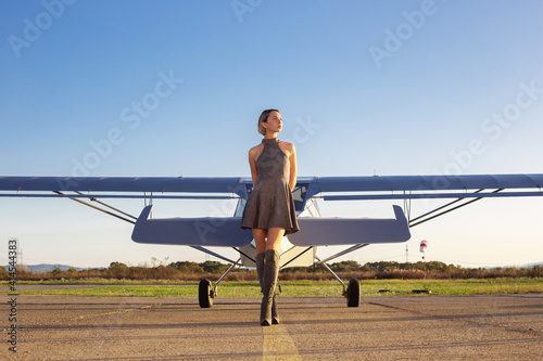 Beautiful woman in gray dress standing in front of private plane. Blue aircraft on aerodrome. Pretty model near small aircraft. Small aviation advertisement.