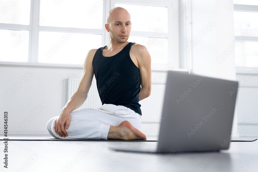 Online training with gadgets. Yogi man with laptop doing yoga, watching training video in daytime in bright studio room, free space. healthy lifestyle concept