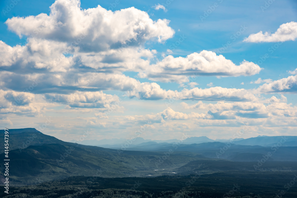 Stunning blue sky day with fluffy, heavenly clouds above rolling mountain peaks during summer time in northern Canada with hazy, mountains in the background.