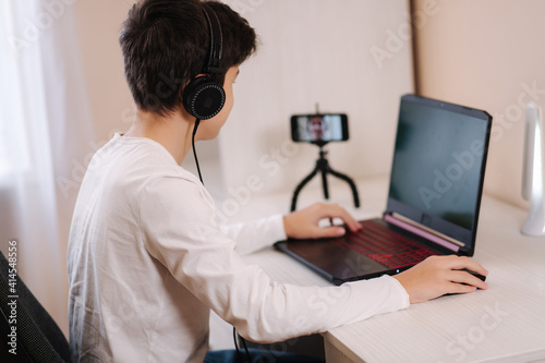 Teenage boy playing game on his pc computer in white room. Gamer capture video on web camera on laptop and phone on tripod