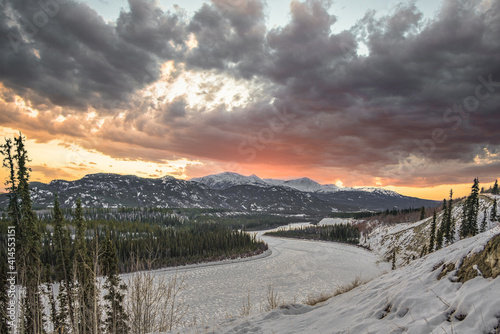 A stunning winter season view of the Yukon River in northern Canada with stormy, snowy looking clouds above at pink, orange sunset hour with snow capped mountains. 