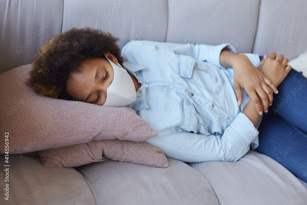 Mixed race teen girl in casual clothes wearing protective face mask relaxing, lying on the couch while spending time at home during quarantine Mixed race teen girl in casual clothes wearing protective face mask relaxing, lying on the couch while spending time at home during quarantine