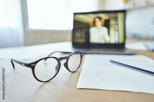 Close up of eyeglasses on the table. Laptop screen with female teacher communicating via video chat app in the background