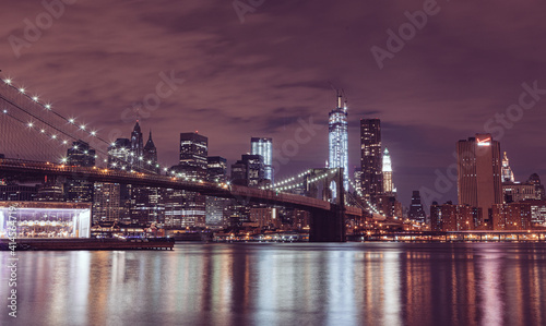 Illuminated Brooklyn Bridge And Buildings By River Against Sky At Night in New York City