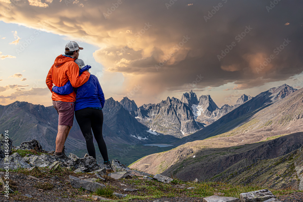 Amazing jagged mountain view in northern Canada, Yukon Tombstone Territorial Park with couple hugging. Orange and purple clothing with stormy looking sky in background. 