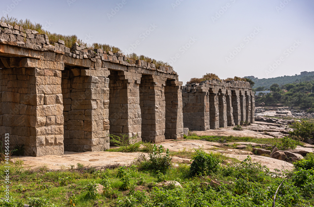 Obraz premium Hampi, Karnataka, India - November 9, 2013: Tungabhadra Bukka Aqueduct brown stone ruins under light blue sky with green weeds and flat surface stones in front.
