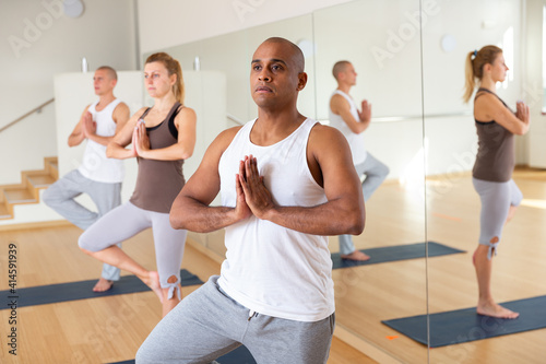 Focused hispanic man standing on one leg in Eka Pada Pranamasana pose during group yoga training in gym