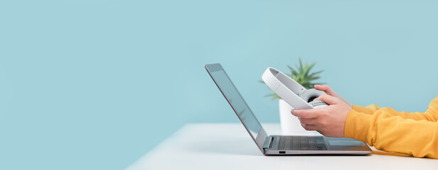 Side view of slim laptop and hands with wireless headphones on grey desk. Blue background. Distant learning. working from home, online courses or support. Audio podcast. vlogger or blogger banner