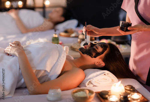 Masseurs hand do holding glass and brushing a facial mask herb black cream to on customer's face as sleep and covered warp by a white on her body