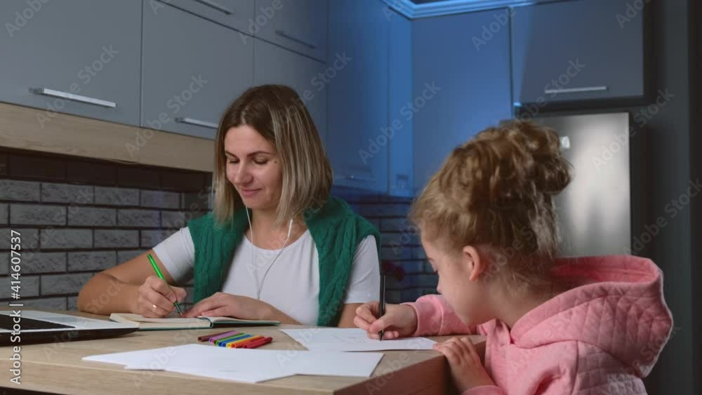 Mother is working in the kitchen at the laptop while her daughter is drawing near her and talking smiling at each other Mother is working in the kitchen at the laptop while her daughter is drawing near her and talking smiling at each other