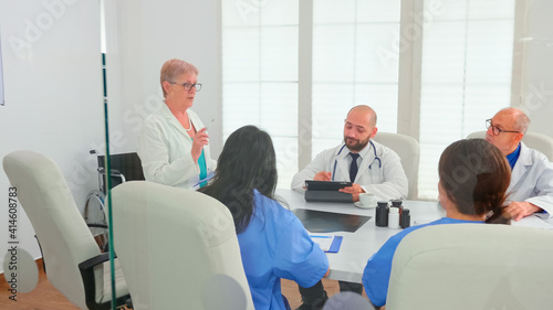 Expert elderly woman doctor having a discussion with medical staff in hospital conference room. Clinic expert therapist talking with colleagues about disease, medicine professional