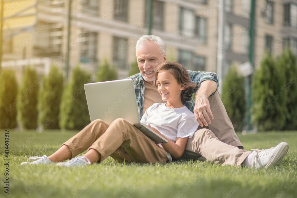 Son and dad sitting together on the grass with a laptop and looking contented Son and dad sitting together on the grass with a laptop and looking contented